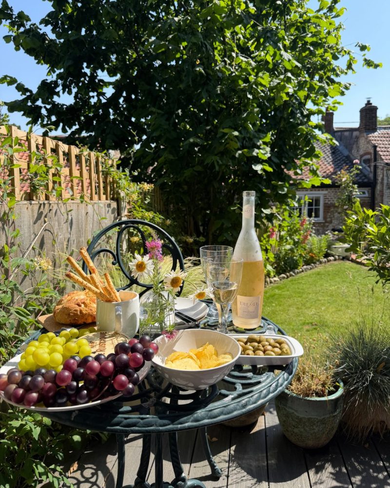 An assortment of picky snacks such as olives, crisps, grapes and breadsticks on an iron round garden table with a bottle of rosé and two glasses. In the background is a beautiful cottage garden in bloom