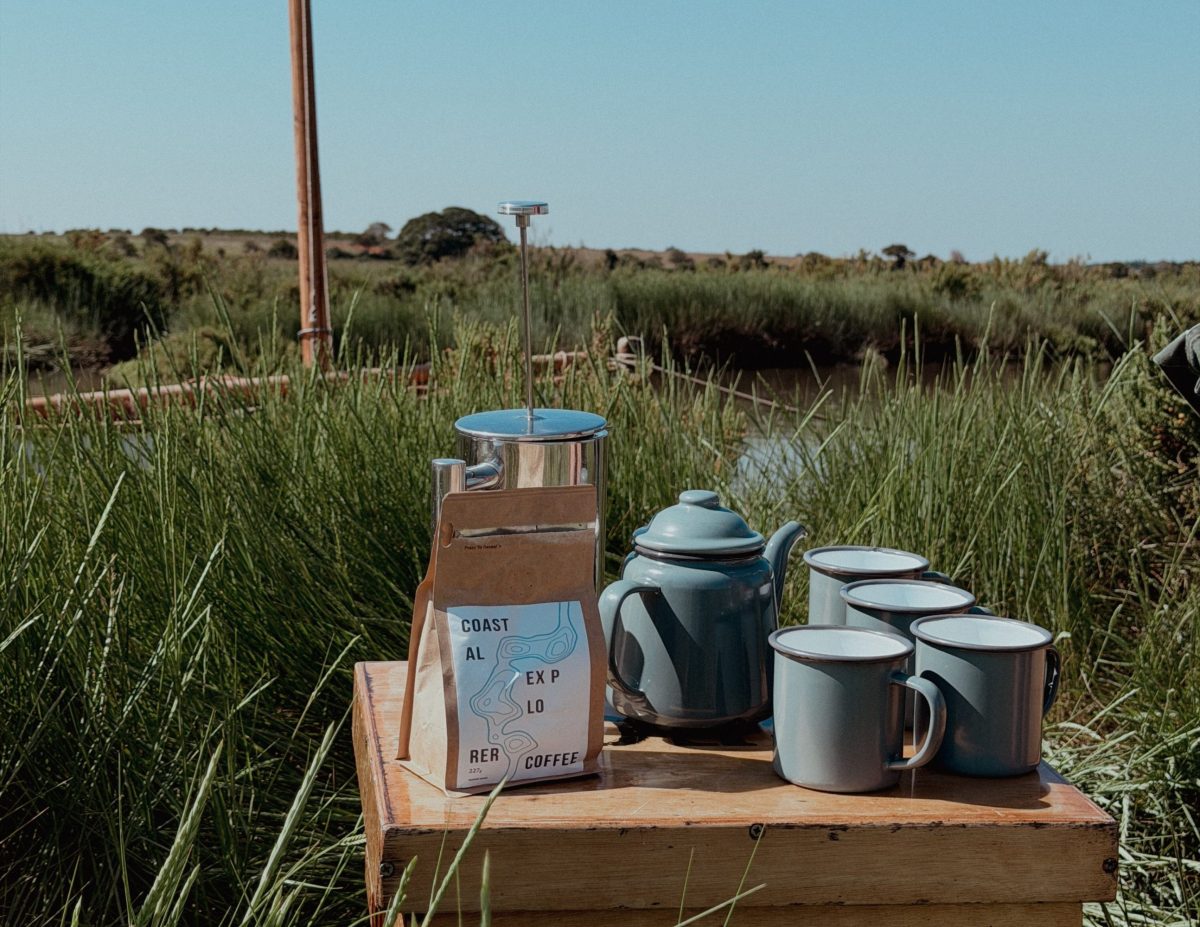 A bag of coffee and ceramic coffee mugs placed on a wooden box. In the background is a boat bobbing amongst the saltmarshes.